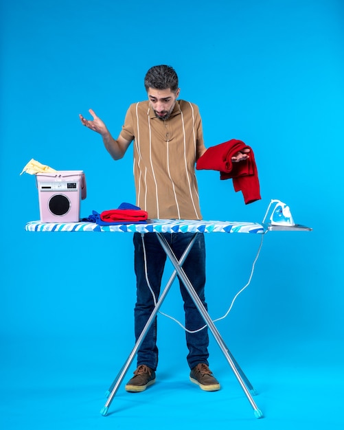 front view young male behind ironing board with clean clothes on a blue background color housework laundry man emotion clean wash machine