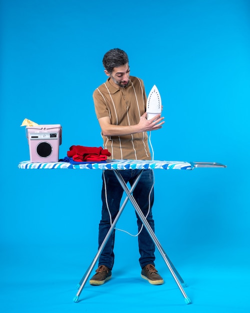 front view young male behind ironing board with towels and iron on the blue background color housework clean laundry man machine wash emotion