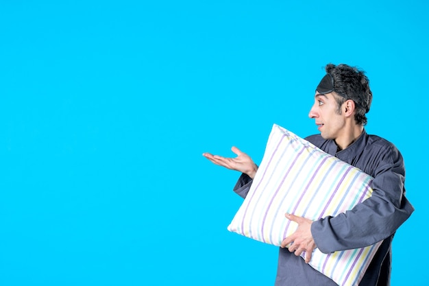 вид спереди молодой мужчина в пижаме держит подушку на синем фоне спальня бессонница мечта темная ночь кровать сон отдых