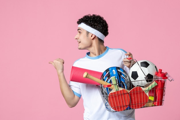 Вид спереди молодой мужчина в спортивной одежде с корзиной, полной спортивных вещей, розовая стена