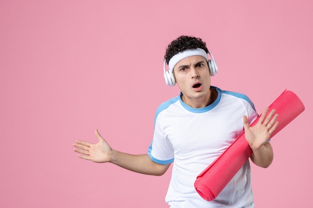 Вид спереди молодой мужчина в спортивной одежде с ковриком для йоги на розовой стене
