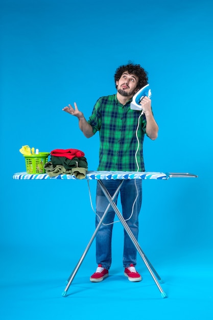 Vista frontale giovane maschio con asse da stiro tenendo il ferro come un telefono su sfondo blu casa vestiti lavatrice lavoro domestico umano clean