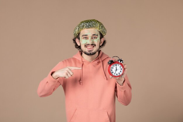 front view young male with mask on his face holding clocks on brown background spa massage skincare beauty human horizontal time skin