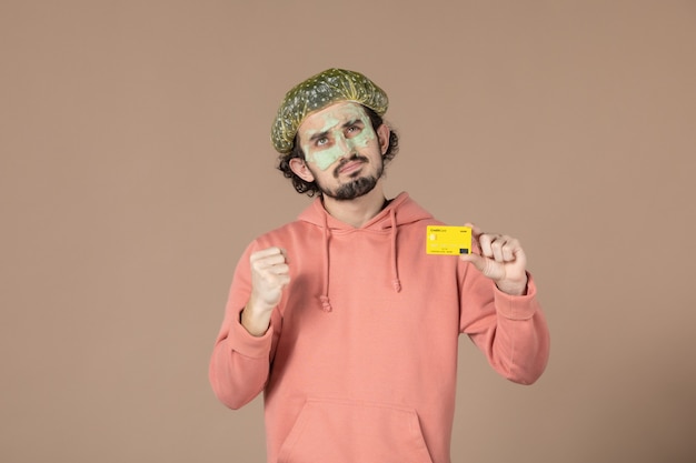 正面図茶色の背景にクレジットカードを保持している彼の顔にマスクを持つ若い男性お金療法スキンケアサロンフェイシャルスキンボディケアスパの色