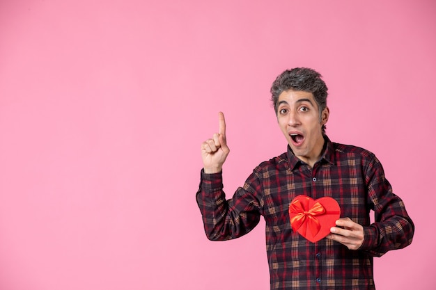 Front view young man holding red heart shaped present on pink wall