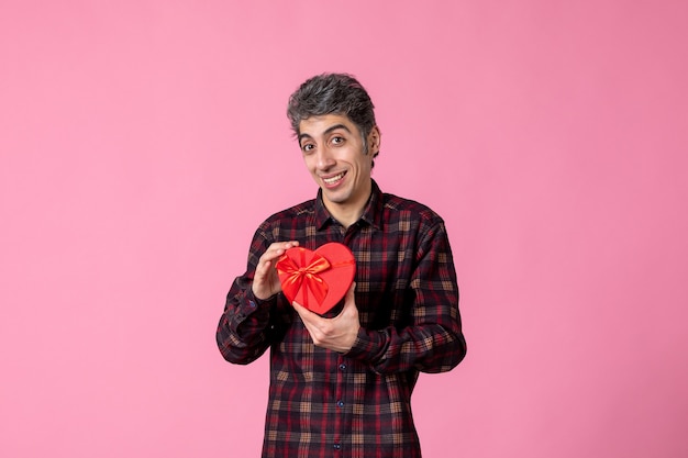 Front view young man holding red heart shaped present on pink wall