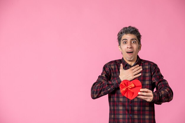 Front view young man holding red heart shaped present on pink wall