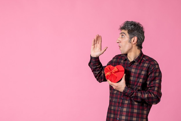 Вид спереди молодой человек, держащий красный подарок в форме сердца на розовой стене