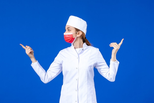 Вид спереди молодая медсестра в медицинском костюме с красной защитной маской на синей стене