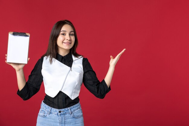 front view young pretty waitress in uniform holding little notepad for orders on red background food color work beauty worker evening job woman