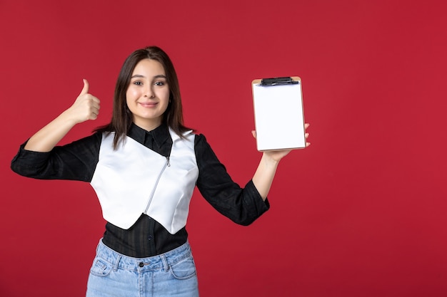 front view young pretty waitress in uniform holding little notepad for orders on red background work food color beauty worker dinner evening job