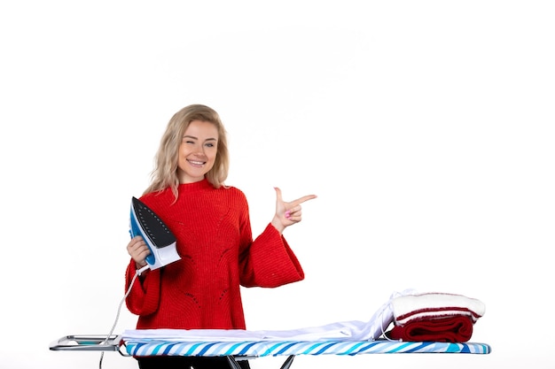 Front view of young smiling beautiful woman holding iron pointing something on the left side on white background