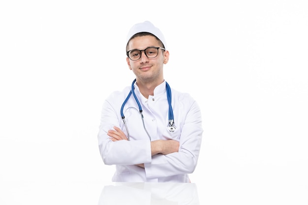 Front view young smiling doctor in medical suit sitting behind desk on white wall