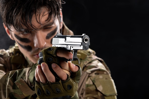 Вид спереди молодого солдата в камуфляже, направленного из пистолета на темную стену