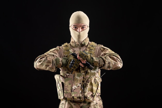 Vista frontale del giovane soldato in uniforme con la pistola sul muro nero