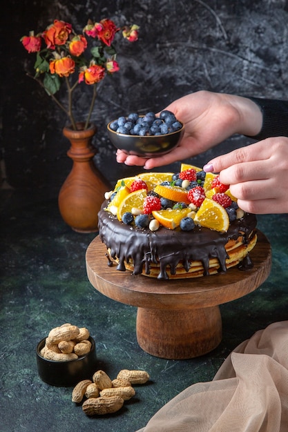 Front view yummy chocolate cake getting decorated with fresh fruits by female on dark wall