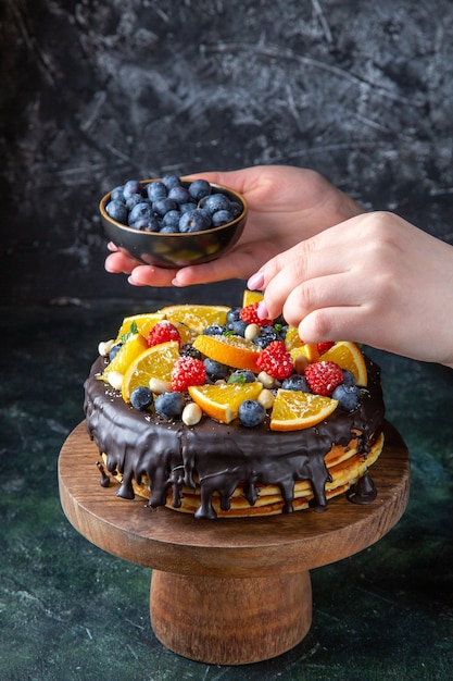 Front view yummy chocolate cake getting decorated with fruits by female on dark wall