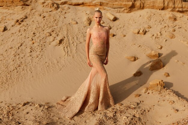 Frontale di una splendida donna bionda in splendido abito d'oro in posa nel deserto, su sfondo di carriera di sabbia.