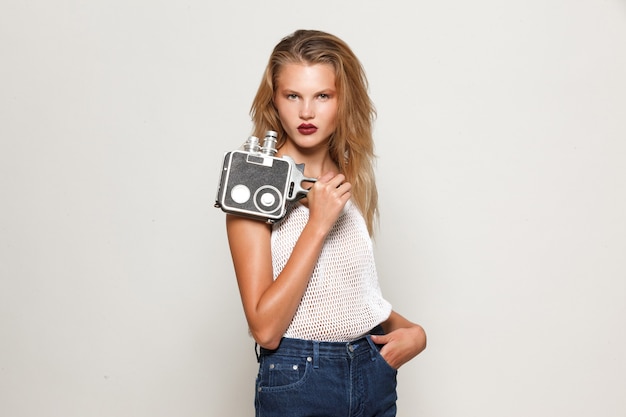 Frontal portrait of girl blonde posing in white fashion clothes , looking at camera, holding a retro camera in hand.