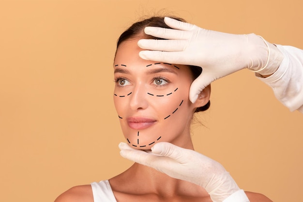 Frontal portrait of young woman getting spa treatment from beautician plastic surgeon making marks on female face