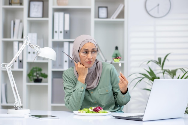 Frontbeeld van een overstuur vrouw die aan een bureau zit met een moderne laptop en een vork vasthoudt om salade te eten