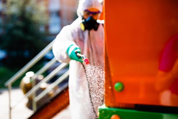 Frontline male worker with face mask and protective workwear\
using pressure sprayer to disinfect children\'s playground. virus\
pandemic protection and prevention concept.