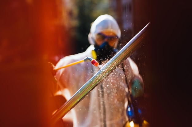 Frontline male worker with face mask and protective workwear using pressure sprayer to disinfect children's playground. Virus pandemic protection and prevention concept.