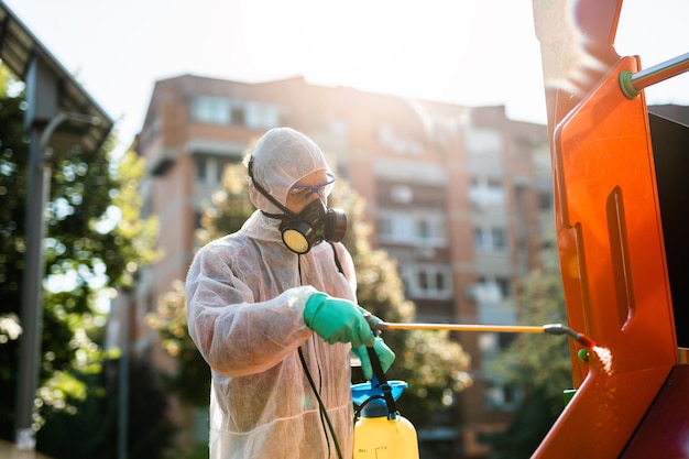 Frontline male worker with face mask and protective workwear\
using pressure sprayer to disinfect children\'s playground. virus\
pandemic protection and prevention concept.