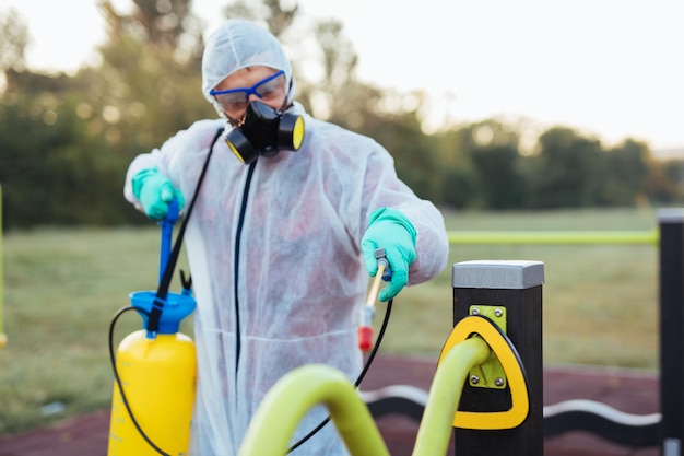 Frontline mannelijke werknemer met gezichtsmasker en beschermende werkkleding met behulp van een drukspuit om de kinderspeelplaats te desinfecteren. Virus pandemie bescherming en preventie concept.