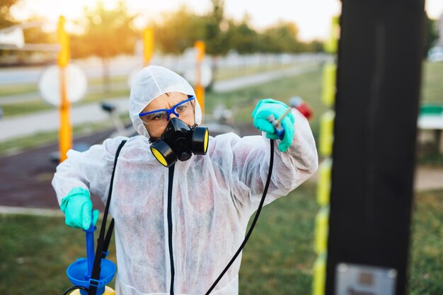 Frontline mannelijke werknemer met gezichtsmasker en beschermende werkkleding met behulp van een drukspuit om de kinderspeelplaats te desinfecteren. Virus pandemie bescherming en preventie concept.