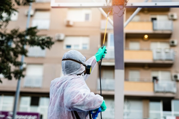 Frontline mannelijke werknemer met gezichtsmasker en beschermende werkkleding met behulp van een drukspuit om de kinderspeelplaats te desinfecteren. Virus pandemie bescherming en preventie concept.