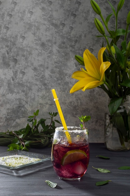 Photo frosty drink with ice and lemon slices on a gray table and a vase with yellow flowers