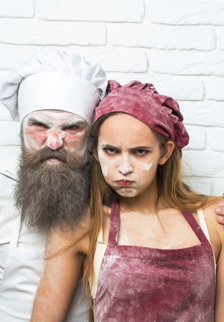 Photo frown handsome man cook or baker with flour on face beard and moustache poses with messy pretty serious girl or beautiful woman on white kitchen wall