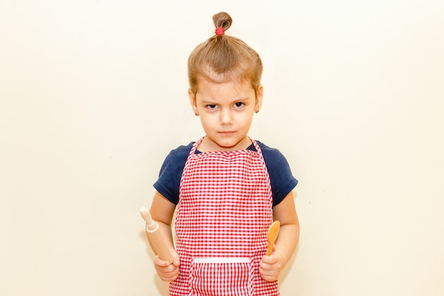 Photo frowned little girl with chef apron holding wooden rolling pin and a spoon on beige background