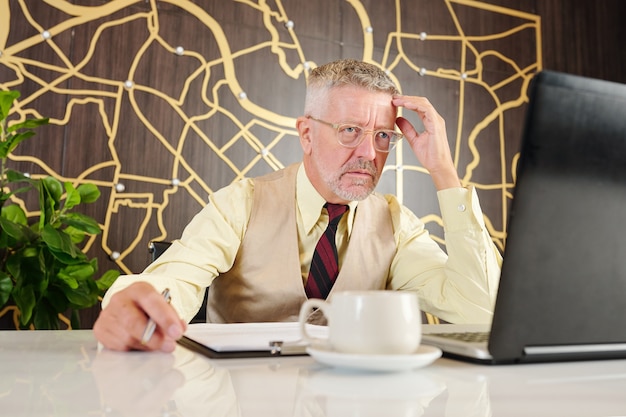 Frowning aged businessman in glasses sitting at table in office and reading e-mail or article on laptop screen