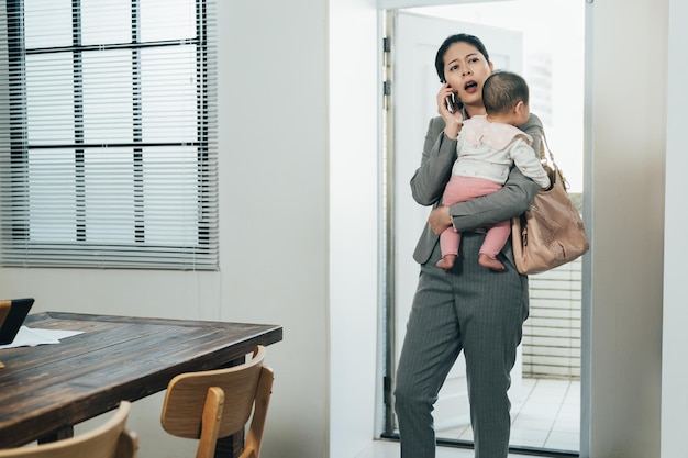 Frowning asian woman ceo entering house holding infant is\
calling her colleague handling trouble at work. busy female manager\
arriving home with kid is nagging at worker through phone for\
mistakes.