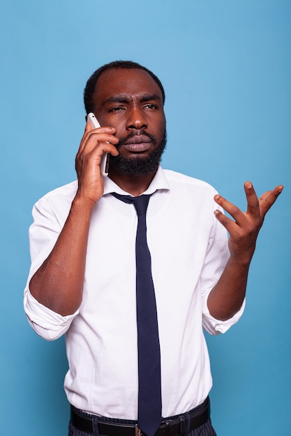 Frowning entrepreneur explaining problem talking on smartphone doing hand gesture on blue background. Upset african american businessman in bad mood trying to understand business conversation.