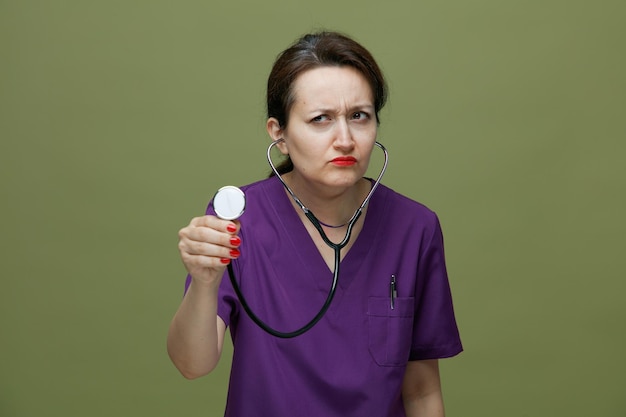 Accigliata dottoressa di mezza età che indossa uniforme e stetoscopio che afferra uno stetoscopio guardando lo stetoscopio di allungamento laterale isolato su sfondo verde oliva