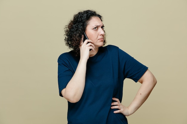 Accigliata donna di mezza età che indossa una maglietta tenendo la mano sulla vita guardando di lato mentre parla al telefono isolato su sfondo verde oliva