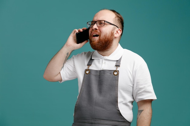 Accigliato giovane barbiere che indossa uniforme e occhiali guardando di lato mentre parla al telefono isolato su sfondo blu