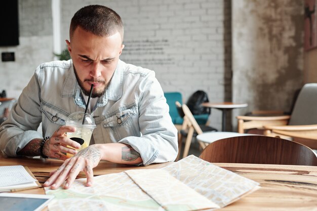 Accigliato giovane uomo barbuto in giacca di jeans seduto in un bar e bere caffè freddo mentre si trovano i posti migliori da visitare utilizzando la mappa