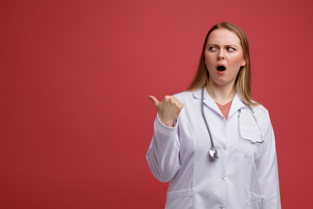 Frowning young blonde female doctor wearing medical robe looking and pointing at side 