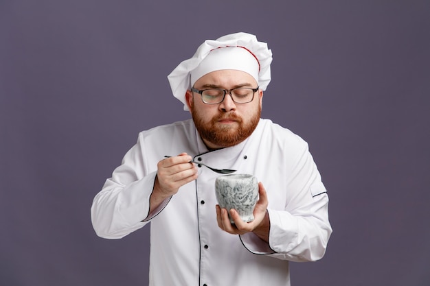 Accigliato giovane chef con gli occhiali uniforme e cappuccio che tiene cucchiaio e ciotola guardando in basso isolato su sfondo viola