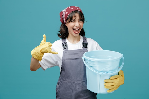 Frowning young female cleaner wearing uniform bandana and rubber gloves holding bucket looking at camera showing hang loose gesture isolated on blue background