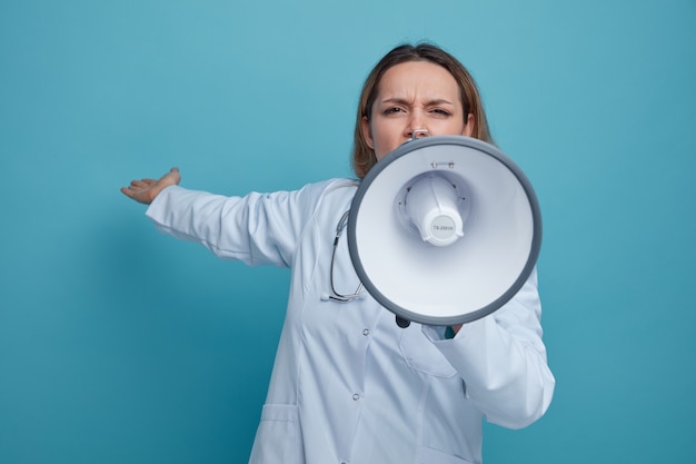 Frowning young female doctor wearing medical robe and stethoscope around neck talking by speaker pointing behind with hand 