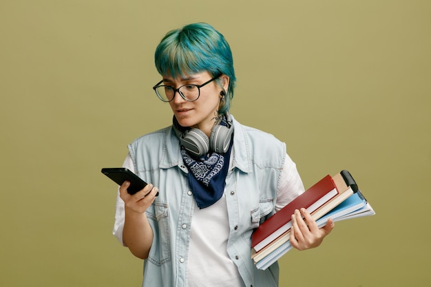 Хмурая молодая студентка в очках, бандане и наушниках на шее, держа блокноты с помощью мобильного телефона, изолированного на оливково-зеленом фоне