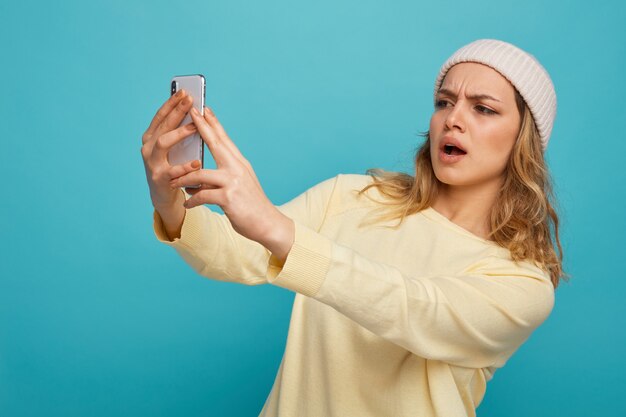 Frowning young girl wearing winter hat stretching out and looking at mobile phone 