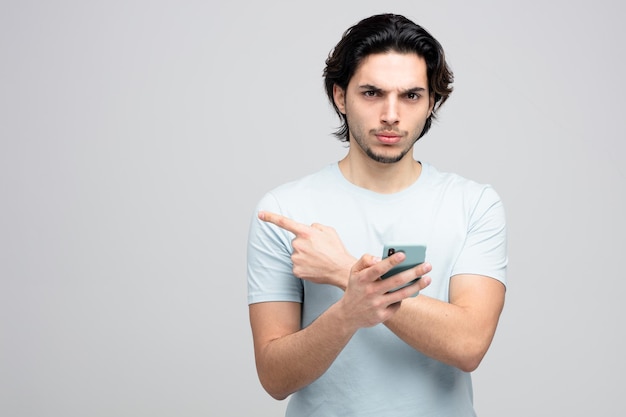 frowning young handsome man holding mobile phone looking at camera pointing to side isolated on white background with copy space