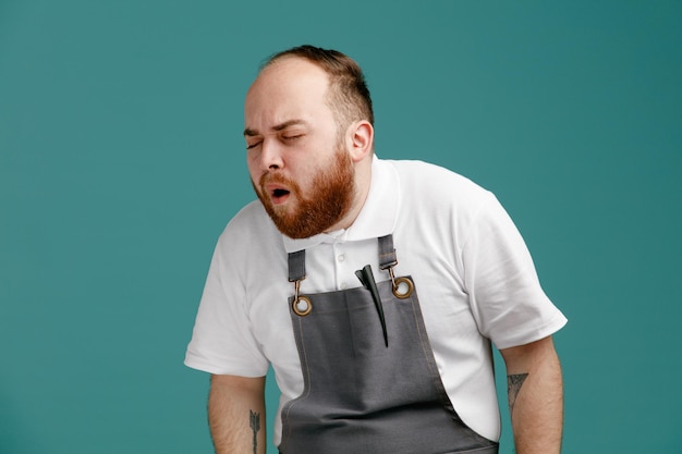 Frowning young male barber wearing white shirt and barber apron getting ready to sneeze with closed eyes isolated on blue background