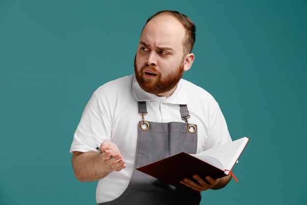 Frowning young male barber wearing white shirt and barber apron holding note pad and pen looking at side isolated on blue background